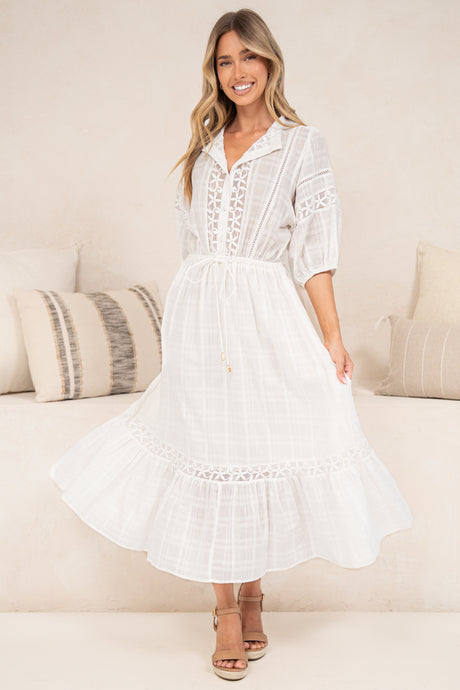 A woman modeling a white midi dress with lace-up front, short sleeves, and a tiered skirt, standing in a bright indoor setting with neutral-toned pillows and a sofa.