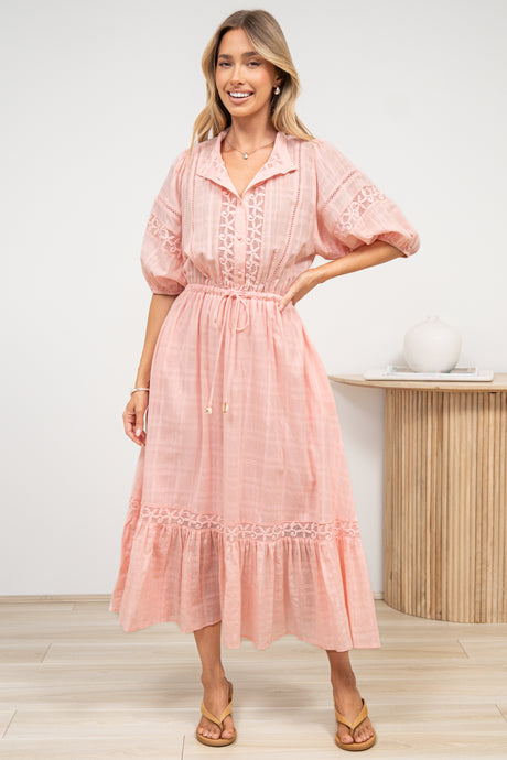 A woman in a romantic pink maxi dress featuring a sheer plaid texture, lace detailing, and flowing fluted hem, posed against an off-white painted wall.