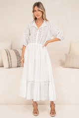 A woman in a flowing white maxi dress featuring sheer plaid-textured fabric, delicate lace detailing, and puff sleeves, posed beside a cream couch.
