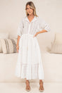 A woman in a flowing white maxi dress featuring sheer plaid-textured fabric, delicate lace detailing, and puff sleeves, posed beside a cream couch.