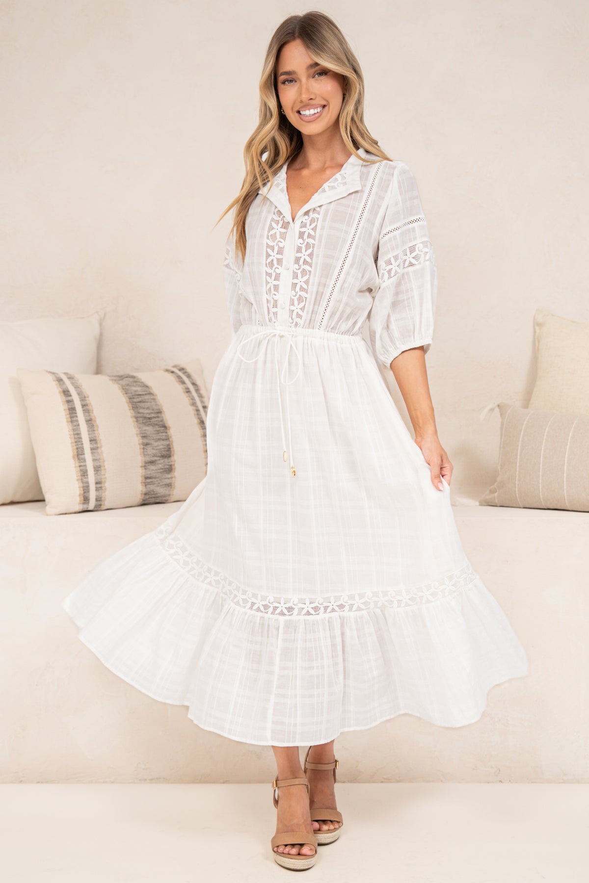 A woman modeling a white midi dress with lace-up front, short sleeves, and a tiered skirt, standing in a bright indoor setting with neutral-toned pillows and a sofa.