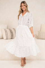 A woman modeling a white midi dress with lace-up front, short sleeves, and a tiered skirt, standing in a bright indoor setting with neutral-toned pillows and a sofa.