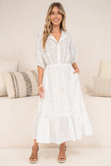 A woman in a flowing white maxi dress featuring sheer plaid-textured fabric, delicate lace detailing, and puff sleeves, posed beside a cream couch.