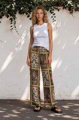 Woman wearing a white tank top and patterned pants standing against a white wall with greenery.