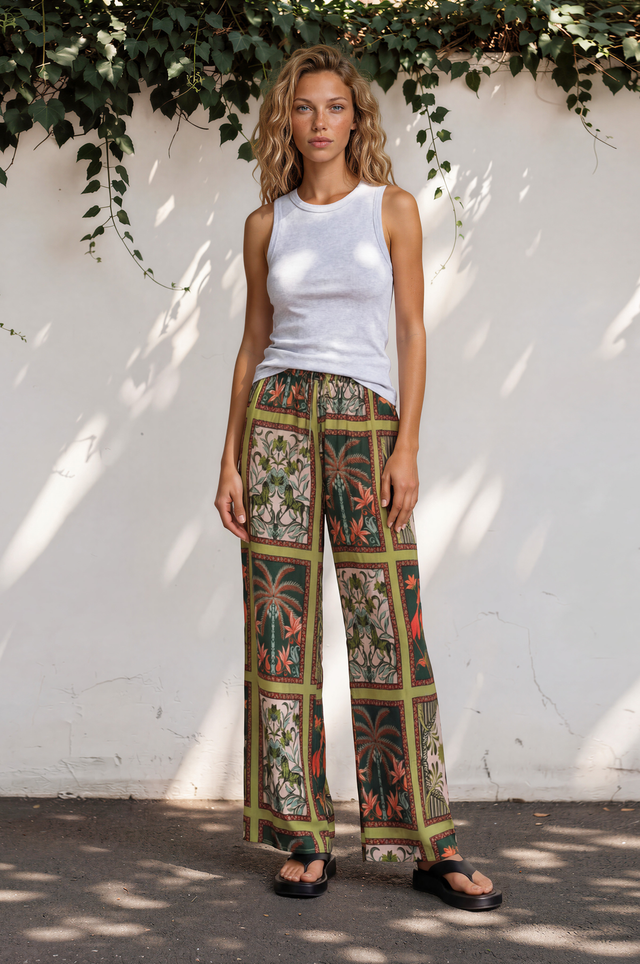 Woman wearing a white tank top and patterned pants standing against a white wall with greenery.