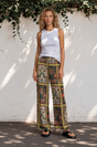 Woman wearing a white tank top and patterned pants standing against a white wall with greenery.