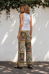 Woman wearing a white top and patterned pants standing against a white wall with greenery.