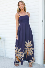 A smiling woman with her arms relaxed wearing the Eira Maxi Dress in Raphael Print, a navy square-neck maxi dress with beaded spaghetti straps, a fitted bodice and palm leaf print hem, by a white wall and doorway
