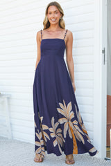 A smiling woman standing against a white wall in the Eira Maxi Dress in Raphael Print, a floor-length navy dress with beaded spaghetti straps, a square neckline and a tan palm leaf print around the hem