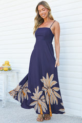 A smiling woman looking down as the skirt of the Eira Maxi Dress in Raphael Print swings out, showing the navy A-line maxi skirt with a tan and orange palm leaf print hem and beaded straps, against a white wall