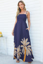 A woman with one hand in her pocket wearing the Eira Maxi Dress in Raphael Print, a navy square-neck maxi dress with beaded spaghetti straps, a smocked bodice and a tan and orange palm leaf print along the hem, against a white wall