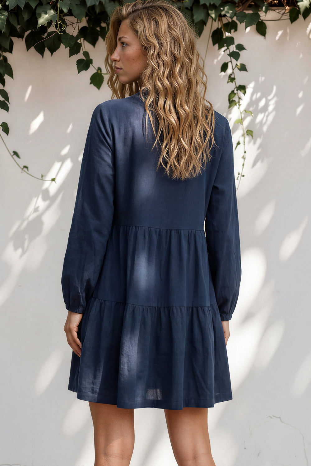 Woman wearing a navy blue dress standing against a white wall with greenery.
