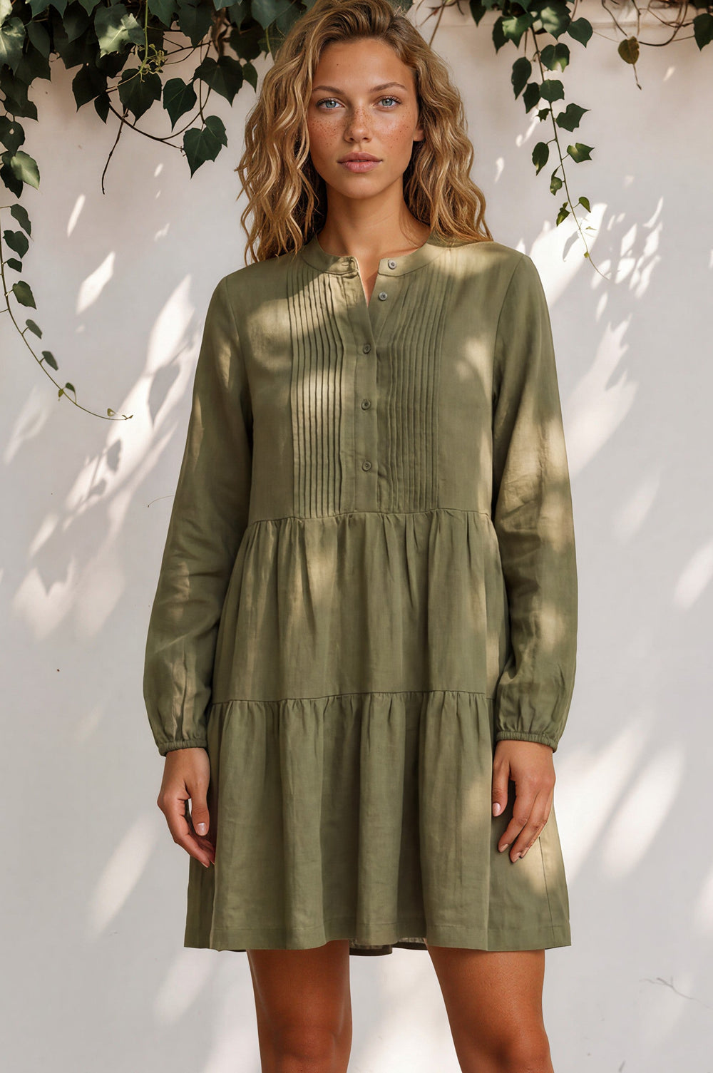 Woman wearing a green dress standing against a white wall with leafy branches.