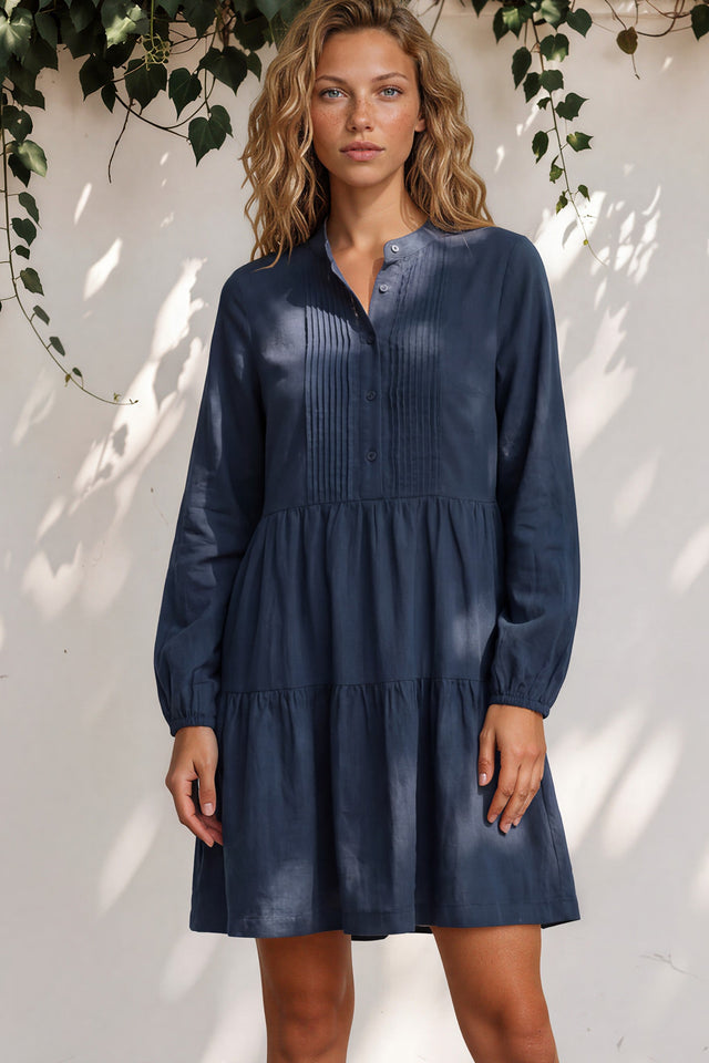 Woman wearing a navy blue dress standing against a white wall with greenery.