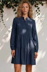 Woman wearing a blue dress standing against a white wall with greenery.