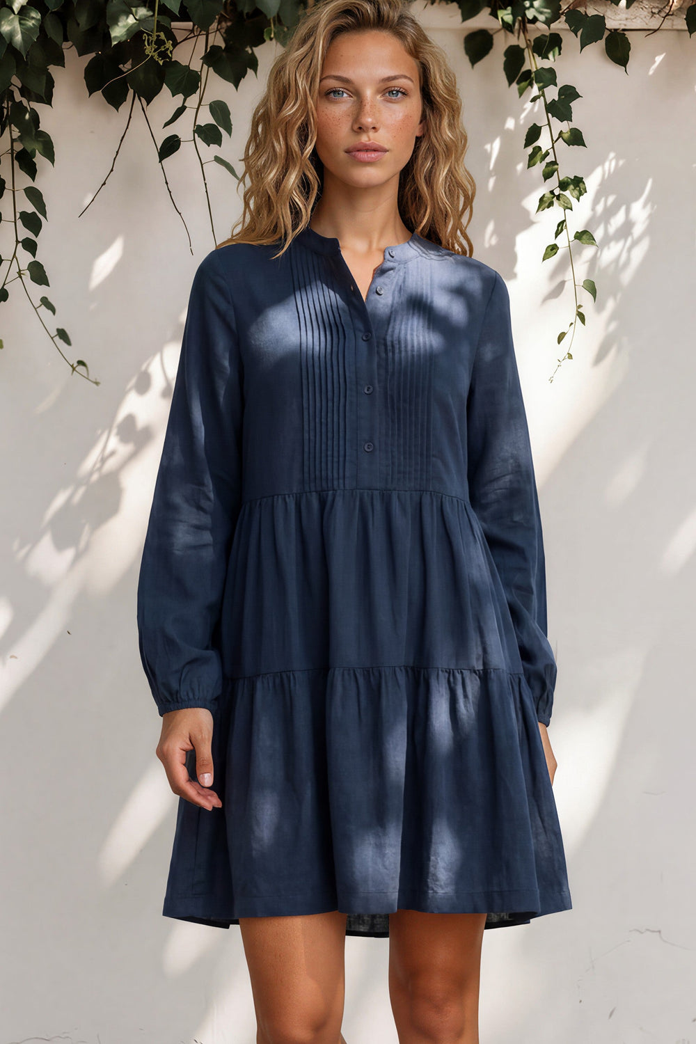 Woman wearing a blue dress standing against a white wall with greenery.