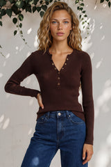 Woman wearing a brown ribbed sweater and blue jeans against a white wall with greenery.