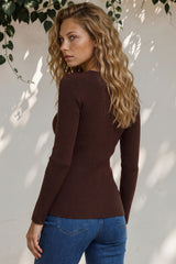 Woman wearing a brown ribbed sweater and blue jeans against a white wall with greenery.