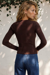 Woman wearing a brown sweater and blue jeans against a white wall with greenery.