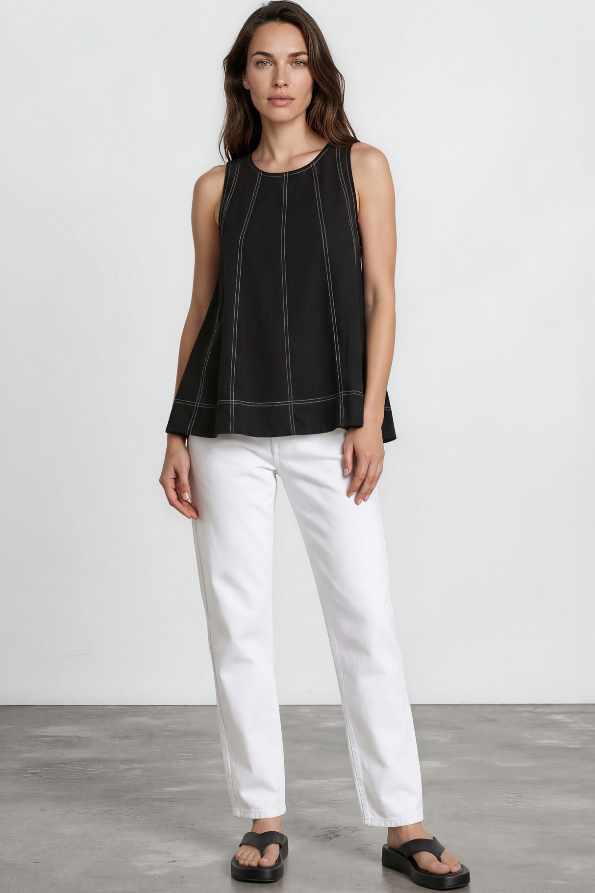 Woman wearing a black sleeveless top and white pants standing against a plain white background