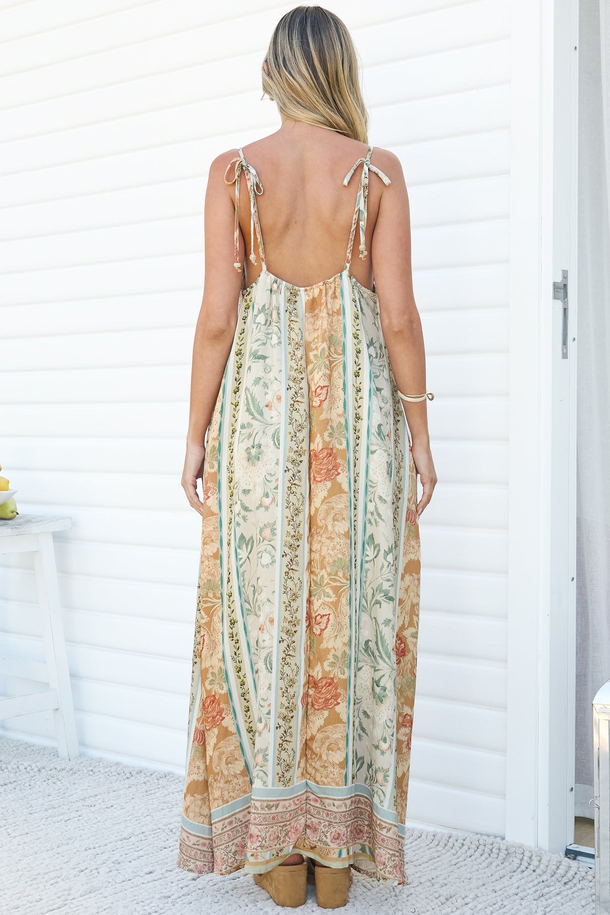 Back view of a woman wearing the Georgette Maxi Dress in Lyla Print, showing the tie shoulder straps, low open back and full floral stripe maxi skirt, against a white wall