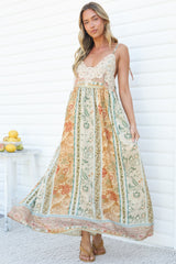 A woman holding out the skirt of the Georgette Maxi Dress in Lyla Print, showing the full, flowing maxi skirt in a cream, sage and orange floral stripe print with a lace motif bodice, against a white wall