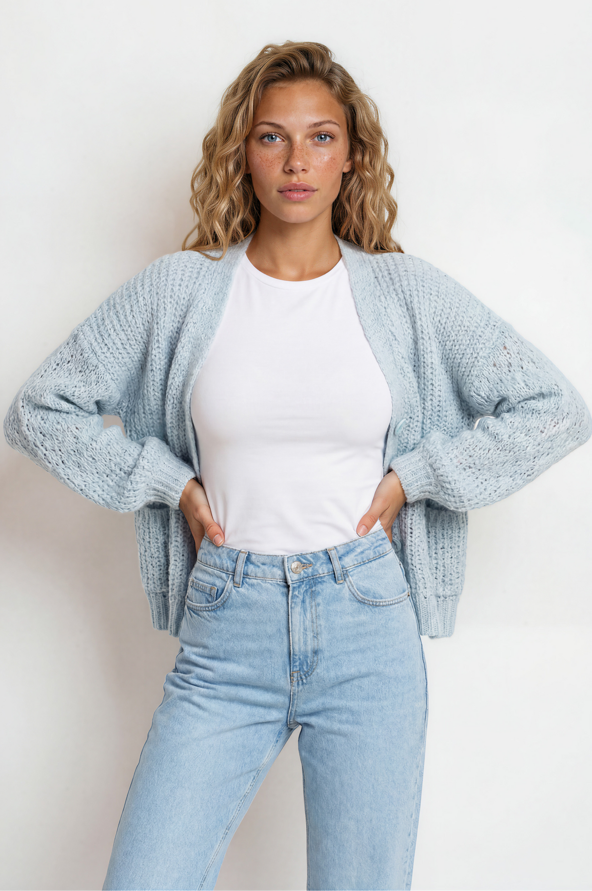 Woman wearing a light blue cardigan, white shirt, and jeans against a plain background