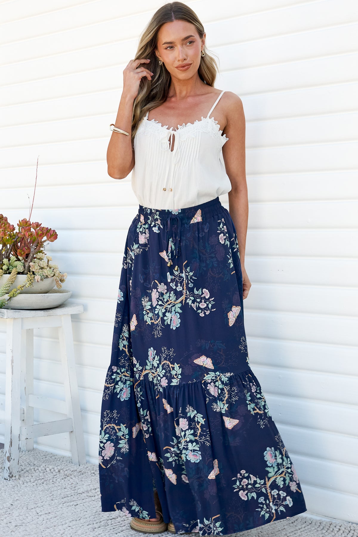 A woman with one hand in her hair wearing the Hellen Maxi Skirt in Colette Print, a high-waisted tiered maxi skirt in a navy floral print with pink and sage blooms, styled with a white lace-trim cami against a white wall