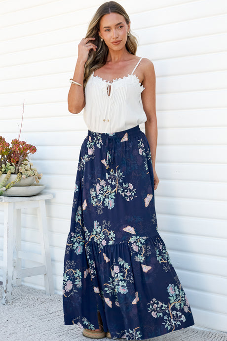 A woman with one hand in her hair wearing the Hellen Maxi Skirt in Colette Print, a high-waisted tiered maxi skirt in a navy floral print with pink and sage blooms, styled with a white lace-trim cami against a white wall