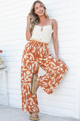A smiling woman leaning against a white wall with one leg bent, wearing the Jaxie Pants in Jaxie Print Rust, flowing wide-leg pants with a paperbag waist and drawstring in a rust and cream swirl print, styled with a white lace-trim cami