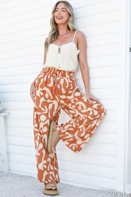 A laughing woman leaning against a white wall with one foot raised, wearing the Jaxie Pants in Jaxie Print Rust, flowy wide-leg pants with a paperbag waist and tassel drawstring in a rust and cream swirl print, with a white cami