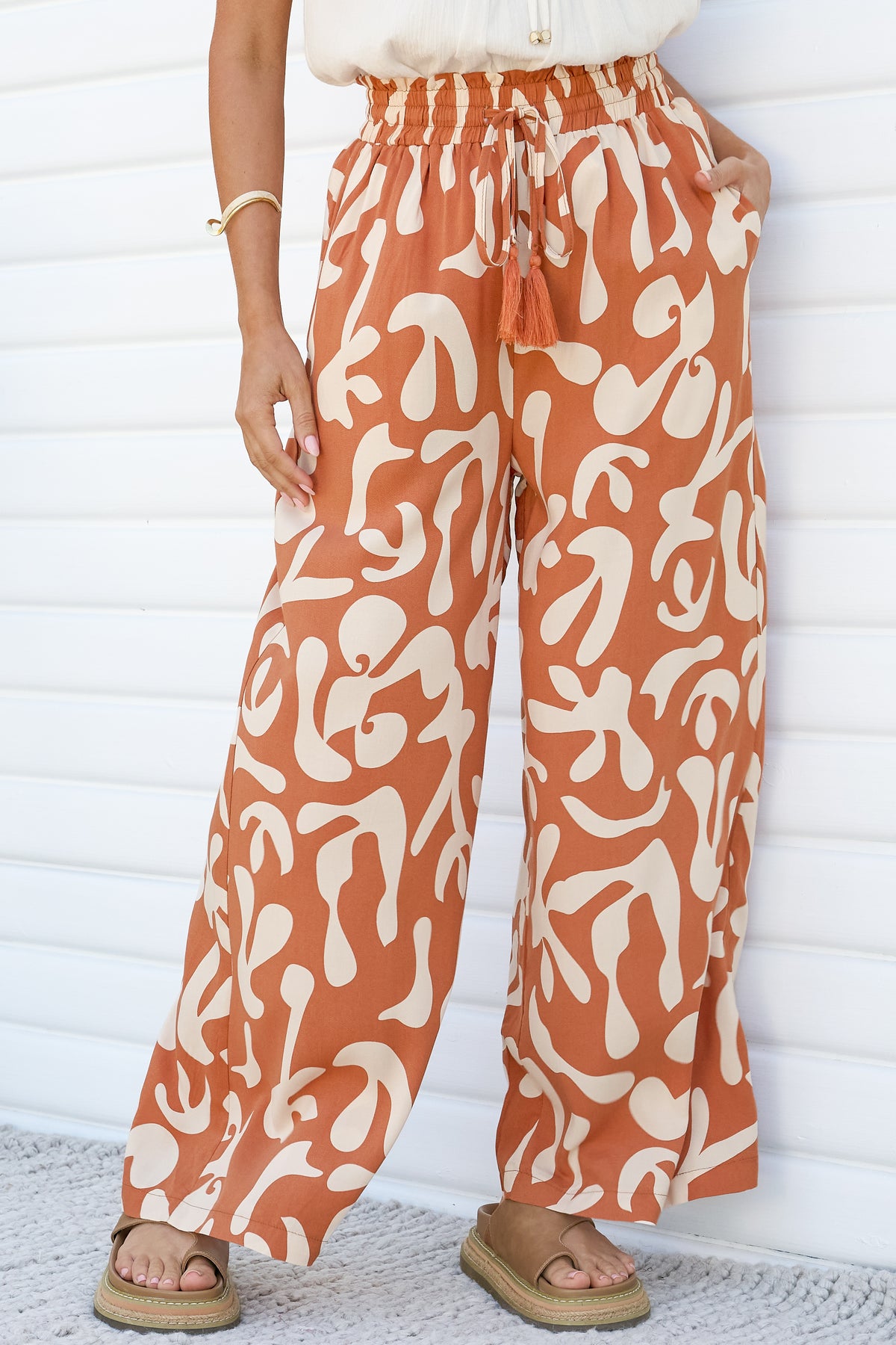 Close-up of the Jaxie Pants in Jaxie Print Rust, showing the smocked paperbag waist, tassel drawstring, side pocket and wide legs in a rust and cream abstract print, worn with platform sandals against a white wall