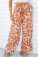 Close-up of the Jaxie Pants in Jaxie Print Rust, showing the smocked paperbag waist, tassel drawstring, side pocket and wide legs in a rust and cream abstract print, worn with platform sandals against a white wall