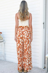Back view of a woman wearing the Jaxie Pants in Jaxie Print Rust, showing the elasticated waist and relaxed wide legs in a rust and cream abstract print, paired with a white cami against a white wall