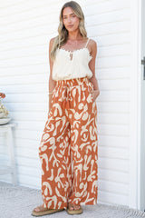 A woman with one hand in her pocket standing against a white wall in the Jaxie Pants in Jaxie Print Rust, relaxed wide-leg pants with a smocked paperbag waist and tassel drawstring in a rust and cream swirl print, with a white cami
