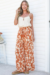 A smiling woman with one hand in her hair wearing the Jaxie Pants in Jaxie Print Rust, wide-leg pants with an elasticated paperbag waist and tassel drawstring in a rust and cream abstract print, paired with a white cami against a white wall