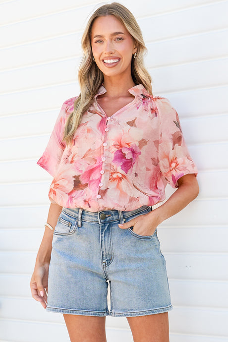 A smiling woman wearing the Jennara Blouse in Marnie Print, a pink and rose watercolor floral button-front blouse with a collar, smocked yoke and short sleeves, tucked into light denim shorts against a white wall