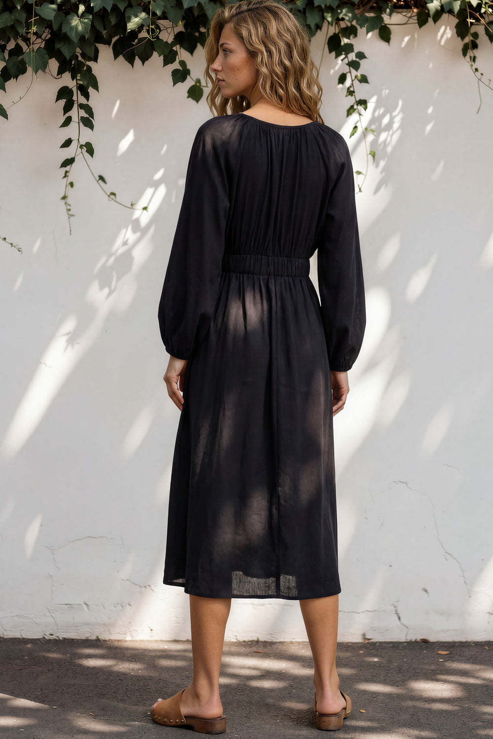 Woman wearing a black dress standing against a white wall with greenery.