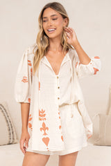 A smiling woman touching her hair while wearing the Joan Blouse in Serenoa Print, a cream tie-front button-down with balloon sleeves and terracotta motifs along the hem, styled with cream shorts in front of a cream sofa