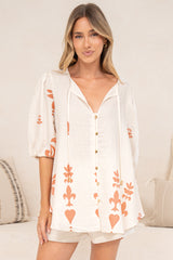 A woman standing front-on in the Joan Blouse in Serenoa Print, a loose cream button-down with a tie-front neckline, balloon sleeves and bold terracotta motifs, worn over cream shorts in front of a cream sofa with neutral cushions