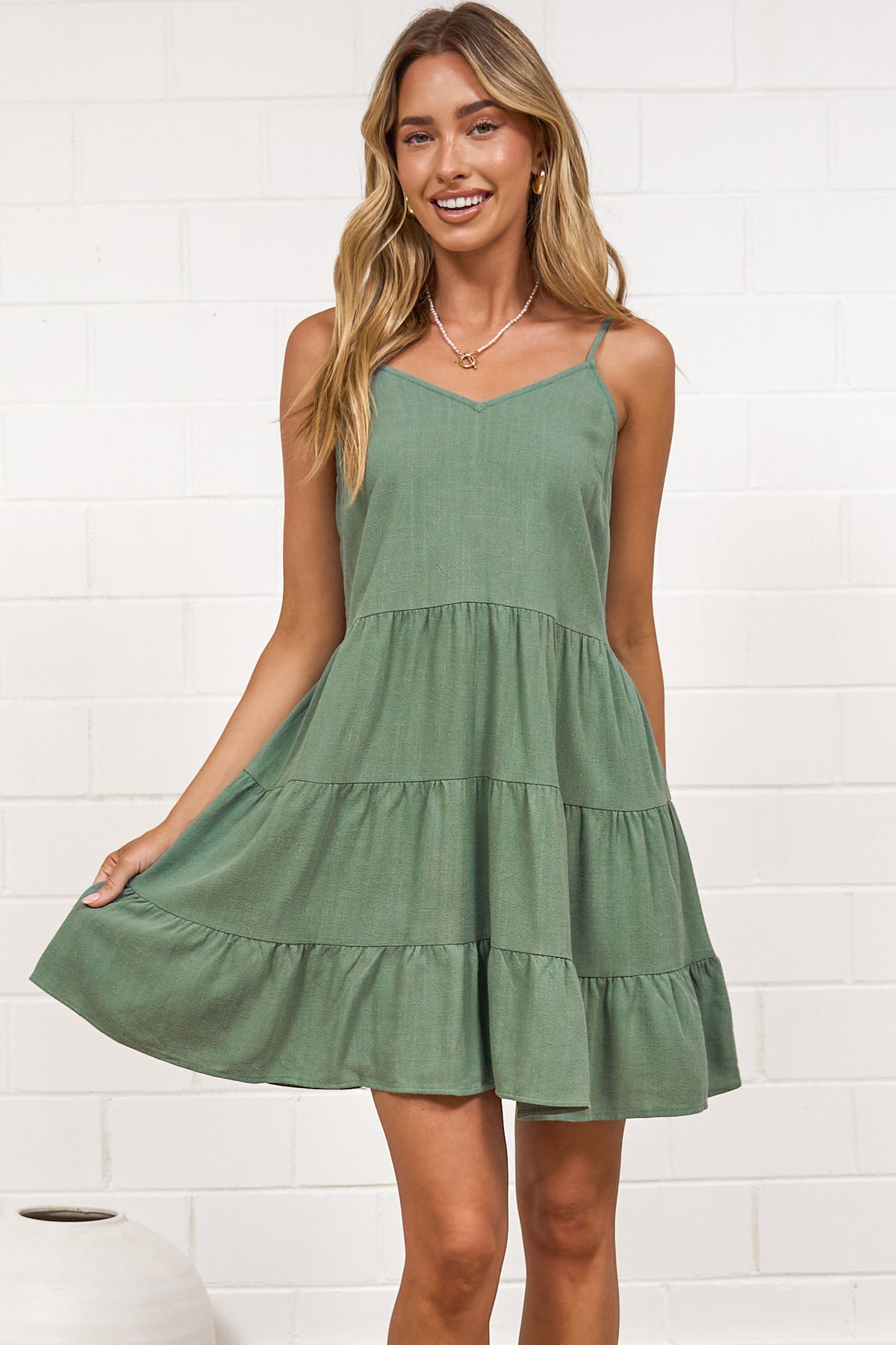 A woman in a dark green mini dress with a V-neckline, adjustable spaghetti straps, and a tiered ruffled hem, posed against a white painted brick wall.