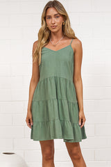 A woman in a dark green mini dress with a V-neckline, adjustable spaghetti straps, and a tiered ruffled hem, posed against a white painted brick wall.