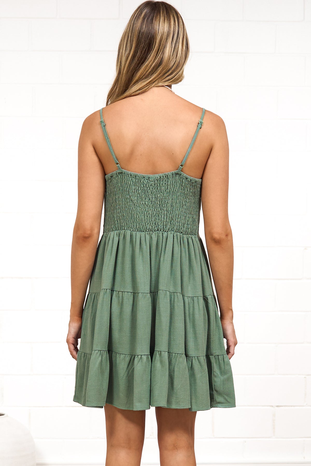 A woman in a dark green mini dress with a V-neckline, adjustable spaghetti straps, and a tiered ruffled hem, posed against a white painted brick wall.