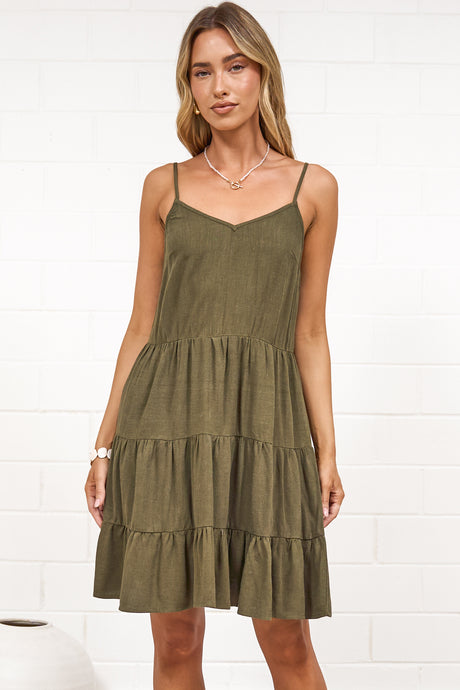 A woman wearing a khaki mini dress with a V-neckline, adjustable spaghetti straps, and a tiered ruffled hem, standing against a white painted brick wall.