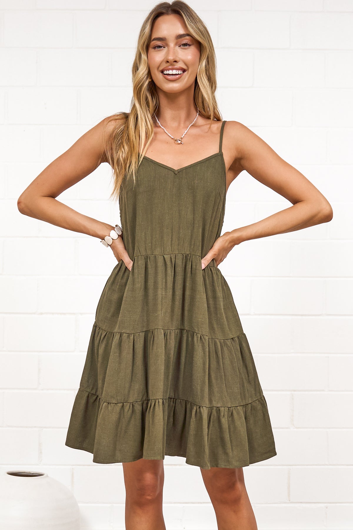A woman wearing a khaki mini dress with a V-neckline, adjustable spaghetti straps, and a tiered ruffled hem, standing against a white painted brick wall.