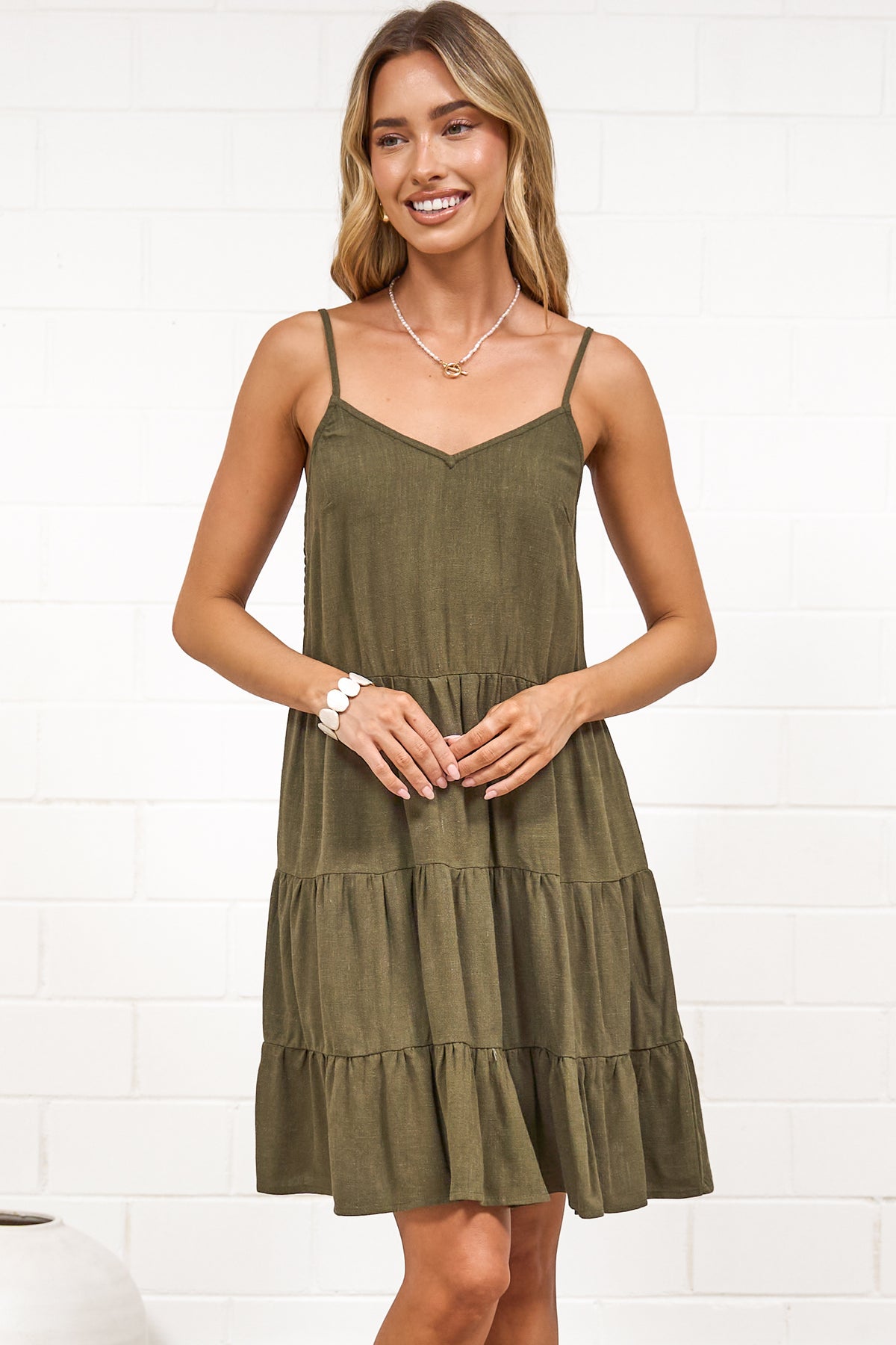 A woman wearing a khaki mini dress with a V-neckline, adjustable spaghetti straps, and a tiered ruffled hem, standing against a white painted brick wall.