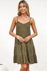 A woman wearing a khaki mini dress with a V-neckline, adjustable spaghetti straps, and a tiered ruffled hem, standing against a white painted brick wall.