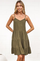A woman wearing a khaki mini dress with a V-neckline, adjustable spaghetti straps, and a tiered ruffled hem, standing against a white painted brick wall.