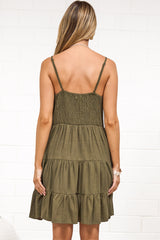 A woman wearing a khaki mini dress with a V-neckline, adjustable spaghetti straps, and a tiered ruffled hem, standing against a white painted brick wall.