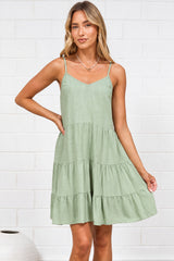 A woman wearing a sage mini dress with adjustable spaghetti straps, a V-neckline, and tiered ruffled hem, standing against a white painted brick wall.